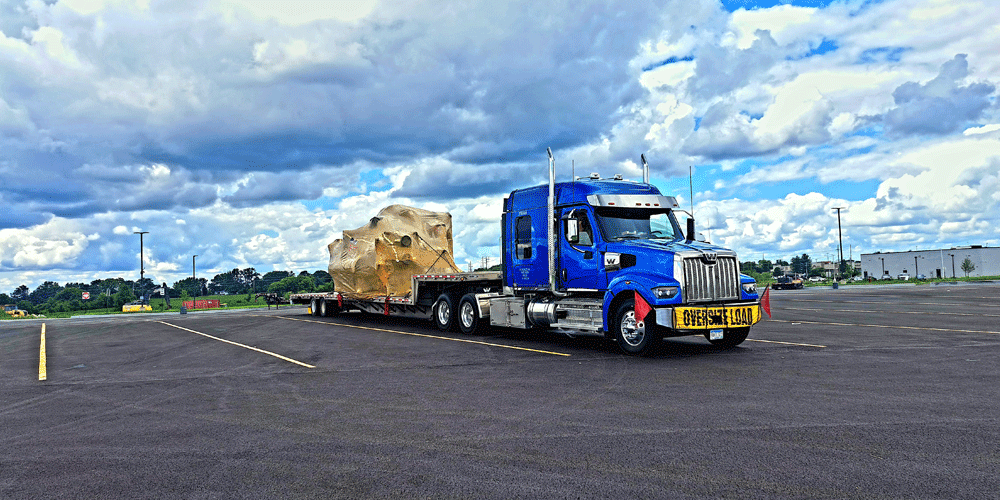 Blue ATS semi-truck with an oversize load banner, red flags on the front, and a tarped load on a stepdeck.