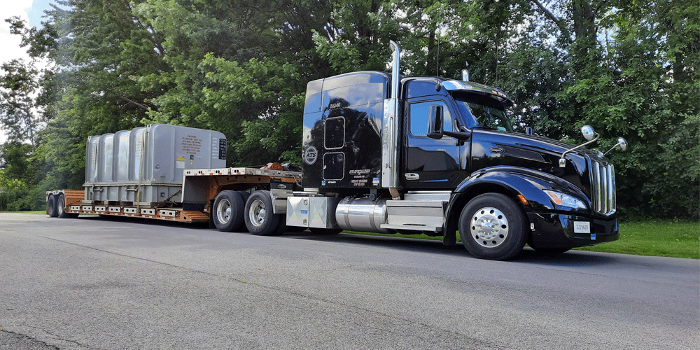 Black semi and stepdeck load parked in front of a heavily wooded area.
