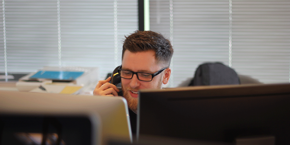 A man on the phone hidden behind two computer screens. He wears black frames.