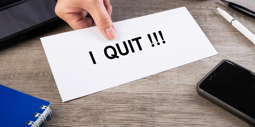White piece of paper that says "I QUIT!!!" Nearby are pens, a smartphone, notebook and laptop.