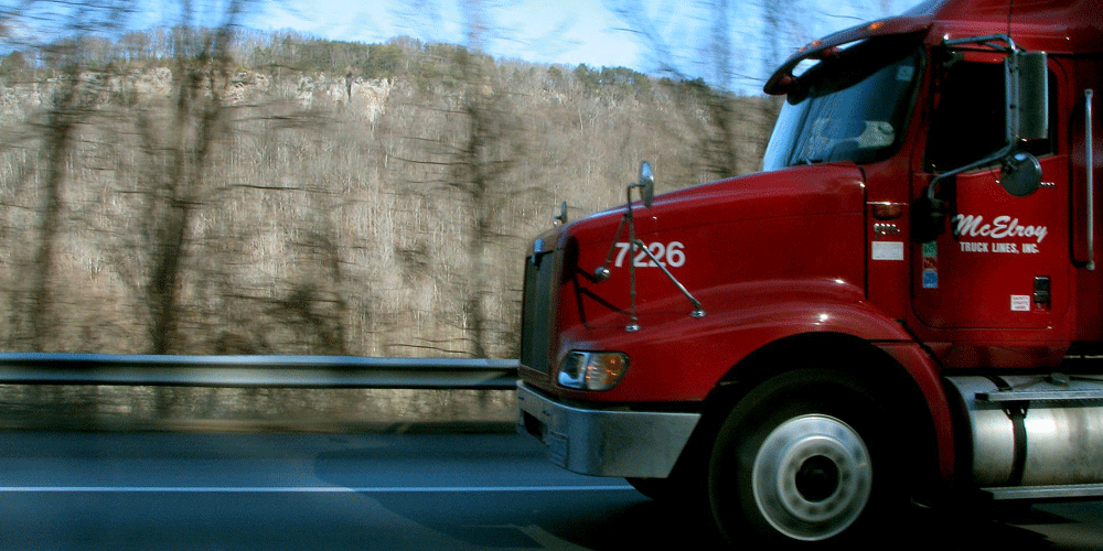 The front glimpse of a McElroy Truck Lines, Inc. red semi-truck as it goes down a wooded road.