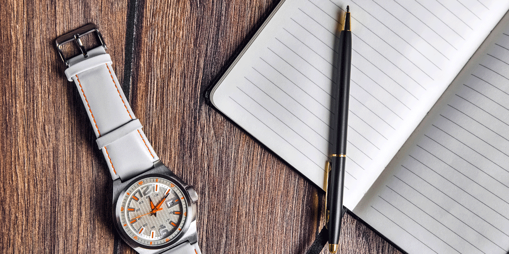 Analog wrist watch laying next to a blank notebook and pen.