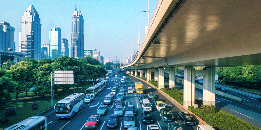 Busy eight-lane road next to an overpass. Tall buildings and skyscrapers are in the background.