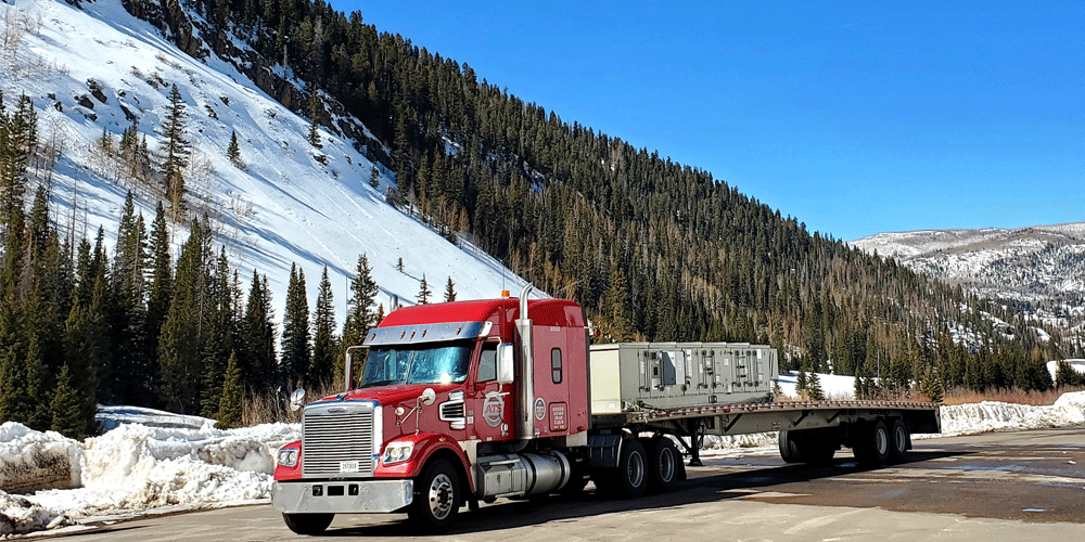 Red tractor and flatbed trailer driving on a snowy mountain road in Colorado.