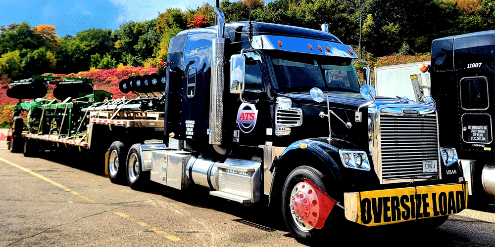 Black truck with oversize load yellow banner and attached stepdeck trailer. It's bright outside and there are bright green and red bushes behind the parked truck.