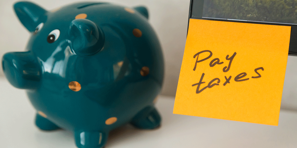 Teal blue piggy bank next to a sticky note reminder that says "pay taxes."