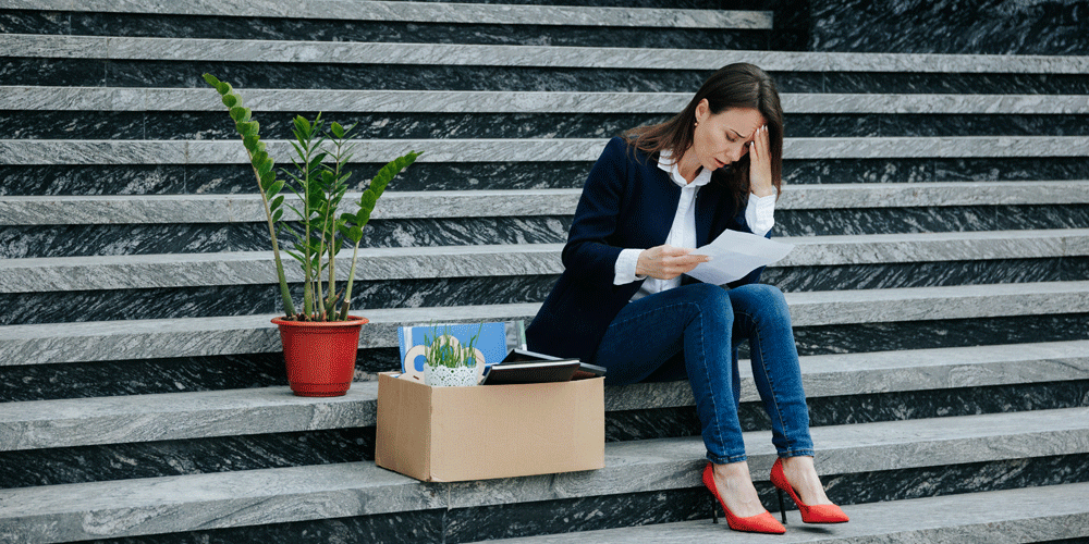 Photo representing layoffs. Woman sitting on concrete steps outside with her head in her hands. She holds a piece of paper and has her desk packed up in a box and a plant next to her.