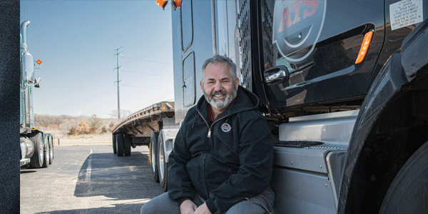 ATS flatbed driver sitting next to his black semi-truck with flatbed trailer; flatbed weight distribution guide for truck drivers; Anderson Trucking Service