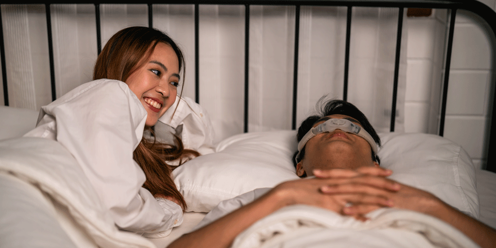 Woman smiling at man in bed as he sleeps soundly with a CPAP machine.