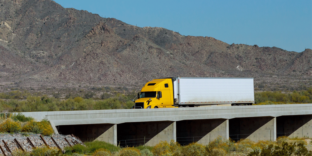 Yellow semi-truck with dry van trailer driving over an overpass. 