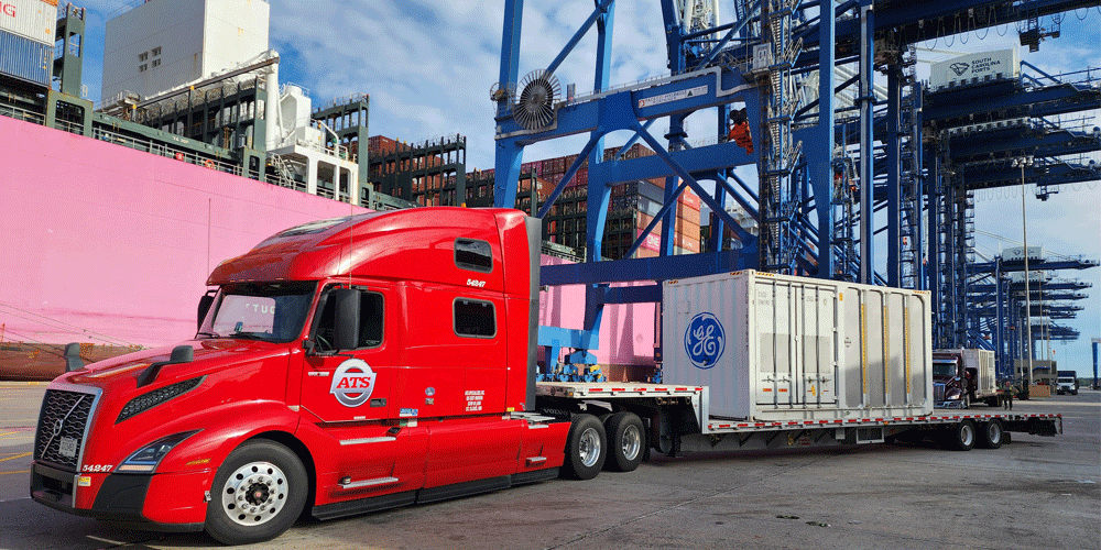 A red ATS tractor at port getting a container loaded onto the flatbed truck.