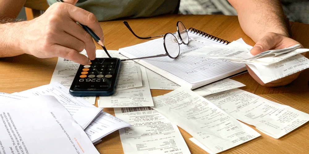 A man sitting at a table with a pile of receipts, notebook, calculator and reading glasses laying in front of him.