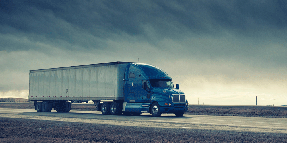 Dry van semi-truck driving with a cloud-covered sky.