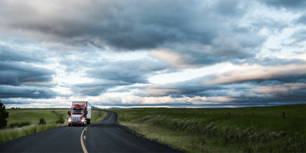 Semi-truck driving a winding road. The sky is full of clouds and it looks like it's dusk.