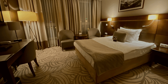 The inside of a hotel room with a bed, two chairs by a curtained window and a desk.