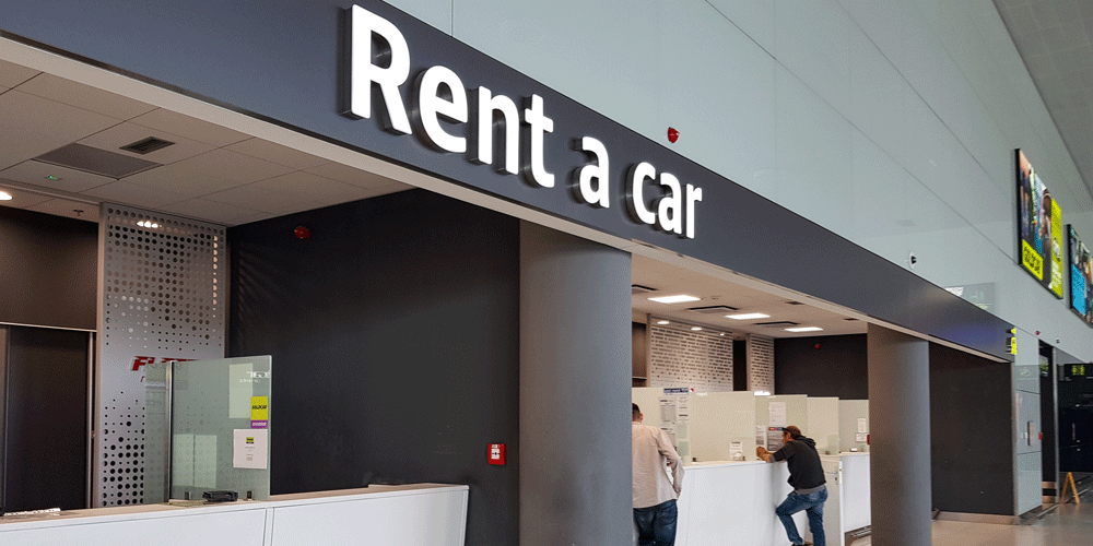 The phrase, "Rent a car" above an entryway to the section to an airport. People stand behind counters under the sign.