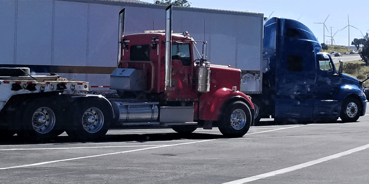 Two big rigs in a parking lot. Wind turbines are seen in the distance. 