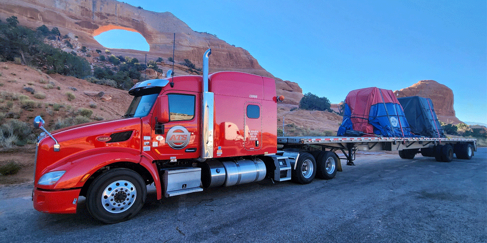 Tarped load on flatbed truck being hauled by a red ATS truck.