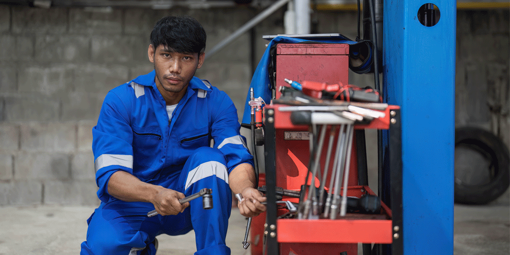 Young mechanic in blue coveralls kneeling down and holding tools.
