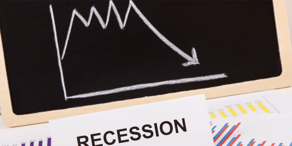 A chalkboard graph showing an arrow pointing down. It lies near pages of graphs and the word "recession" typed in black on white paper.