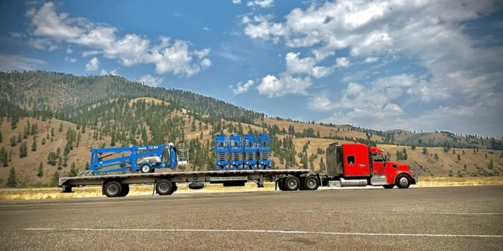 Red truck with a flatbed trailer hauling blue equipment. There are tree-covered hills in the background