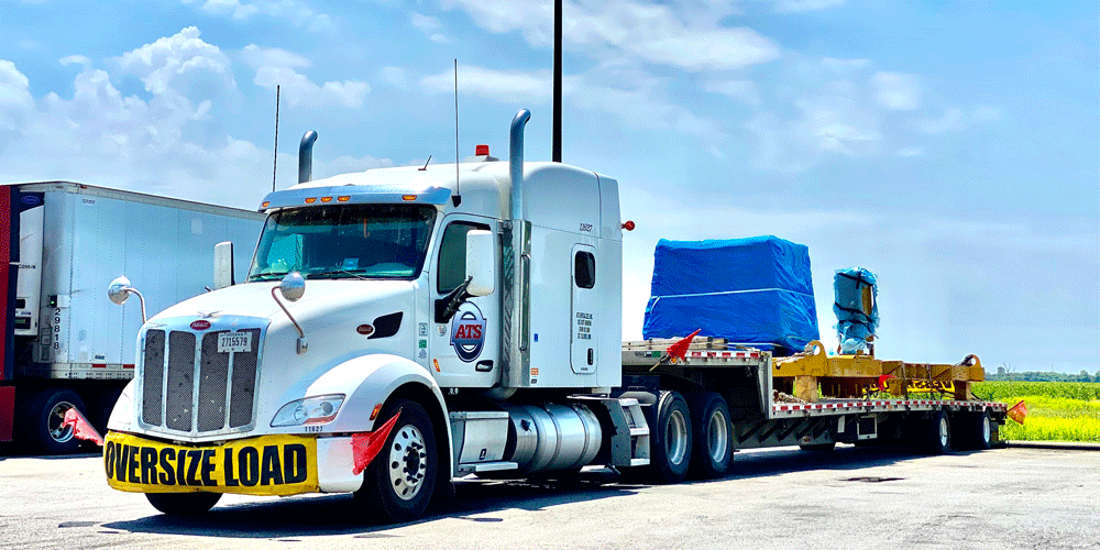 White semi-truck with an "oversize load" yellow banner on the front. There's a flatbed trailer attached.