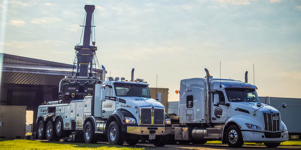 A white truck with crane parked next to a white semi with a flatbed trailer attached.