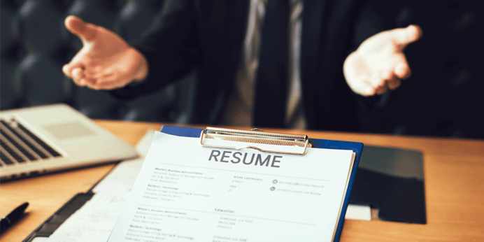 A resume attached to a clipboard. It sits on a desk in front of a person in a suit holding out their arms.