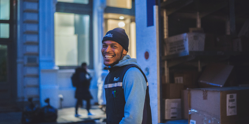 Truck driver in a beanie, a vest, and a sweatshirt. He smiles at the camera.