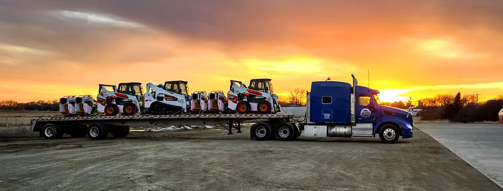 An ATS truck hauling a flatbed trailer loaded with multiple pieces of heavy machinery against a sunset.