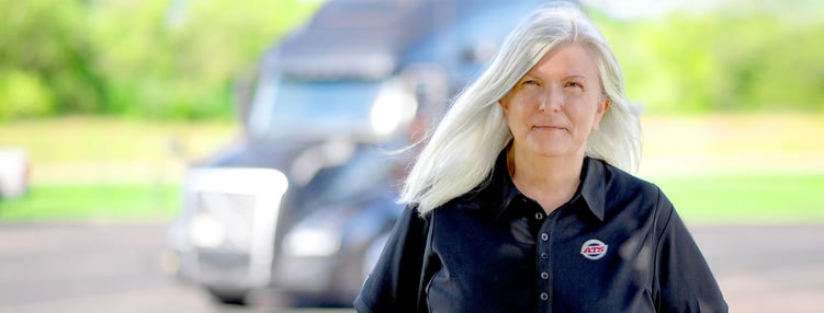 Woman ATS driver stands with out-of-focus semi-truck in background, OTR pay at ATS, Anderson Trucking Service