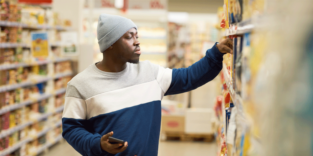 Man in stocking cap and sweater holding his phone while he grocery shops.